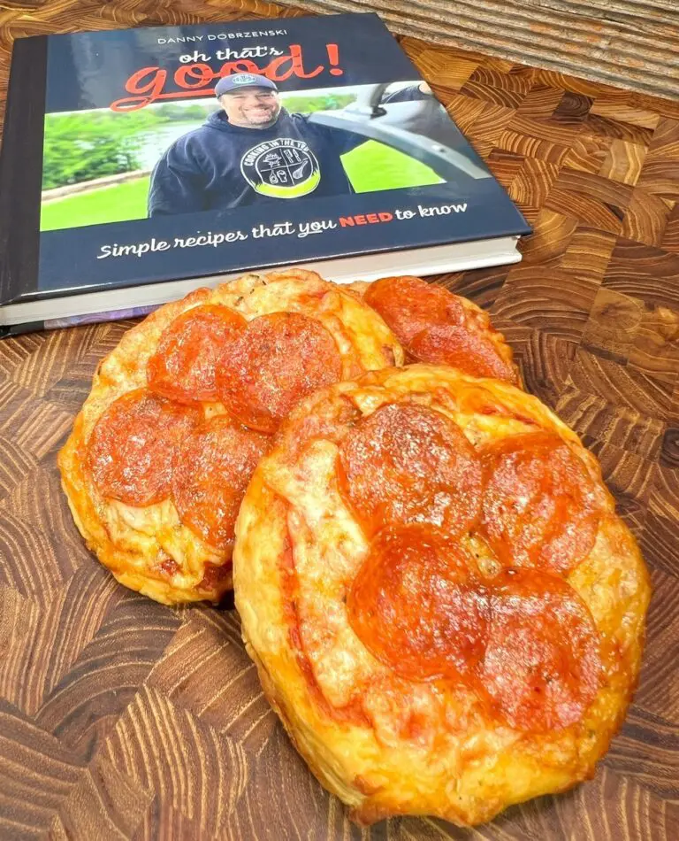 Two stuffed pizza biscuits rest on a wooden surface in front of a cookbook titled "Oh That Good!" The cover showcases a person in a black shirt and cap, smiling warmly, promising delightful recipes within.