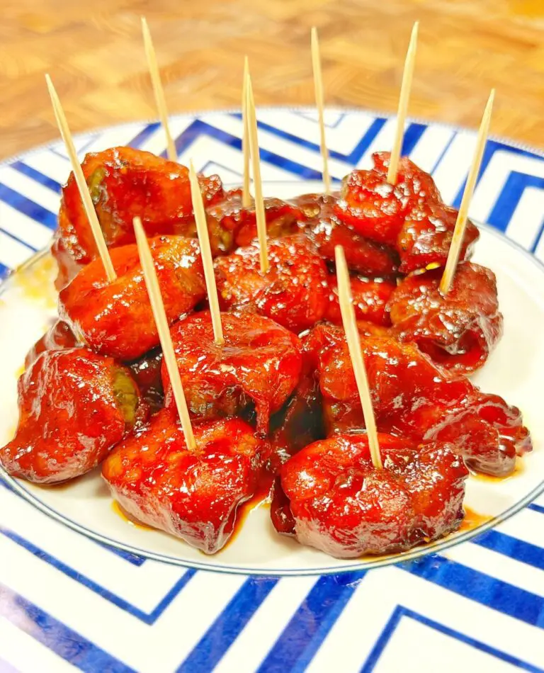 A plate of bacon-wrapped meatballs with toothpicks, arranged on a decorative blue and white plate. The meatballs are covered in a shiny red sauce reminiscent of burnt ends. The background shows a wooden surface.