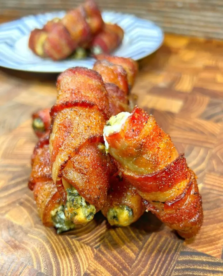 Close-up of crispy spinach-infused bacon-wrapped jalapeño poppers on a wooden surface, with a blurred plate of more poppers in the background. The bacon is golden brown, showcasing the jalapeños and cream cheese filling.