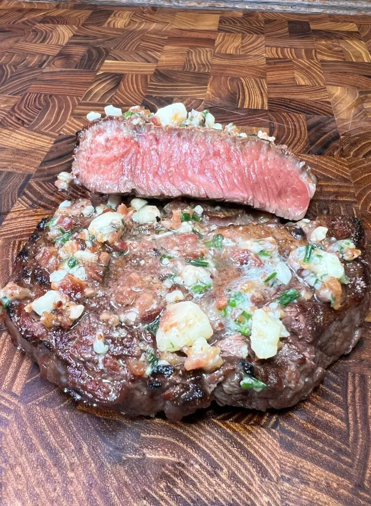 A juicy, medium-rare steak garnished with blue cheese butter sits on a wooden cutting board. A slice of the steak shows its pink, tender interior, highlighting the marbled texture and seasoning.