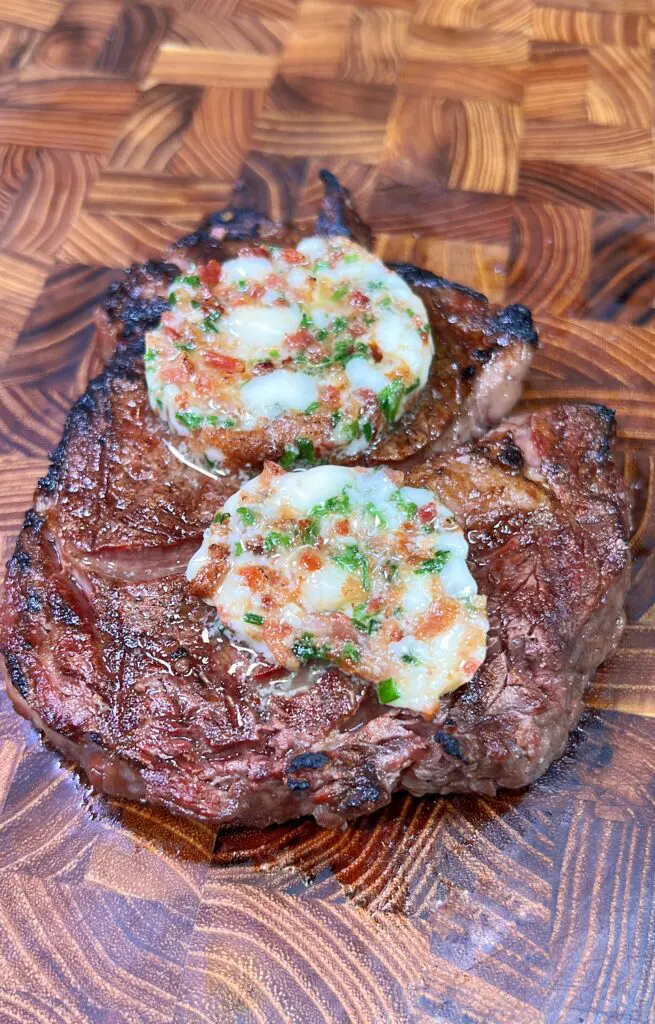 A grilled steak topped with two rounds of blue cheese butter, garnished with chives and bits of bacon, served on a wooden cutting board with a distinct grain pattern.