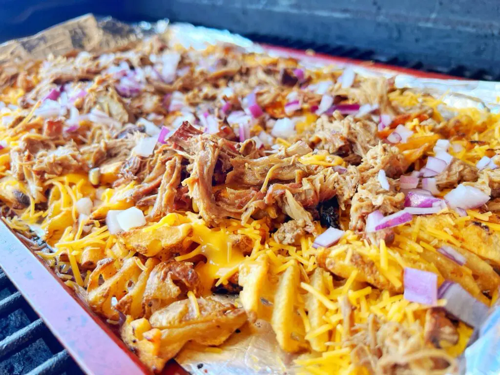 Close-up of loaded fries topped with shredded cheese, pulled pork, and diced red onions, served on aluminum foil. The dish is set on a grill, showcasing its colorful and hearty toppings.
