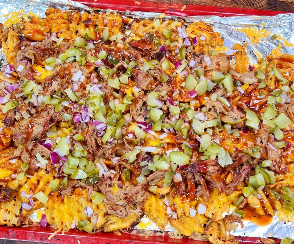 A tray of waffle fries topped with shredded pulled pork, chopped pickles, red onions, melted cheese, and a drizzle of barbecue sauce. The fries are spread over a foil-lined baking sheet.