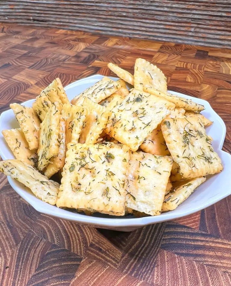 A white bowl brimming with square crackers, reminiscent of a dill pickle snack, is topped with herbs and set on a wooden table with a woven pattern.