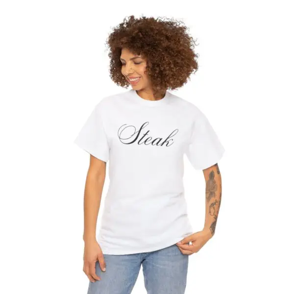 A person with curly hair grins while posing in a white Steak Tee with the word "Steak" elegantly scripted. They’re rocking blue jeans and flaunt tattoos on their left arm, set against a plain white background—a perfect snapshot of style.