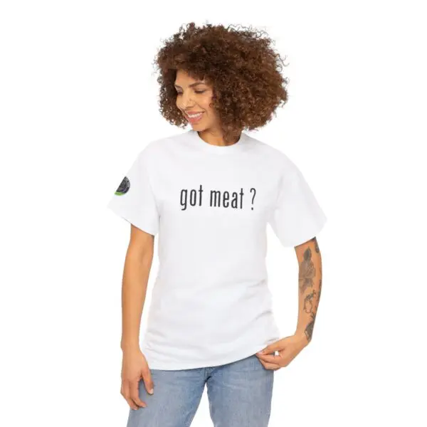 A person with curly hair smiles while wearing a white "got meat ?" T-shirt. Their tattooed arms and blue jeans add to an effortlessly casual vibe, exuding easy confidence.