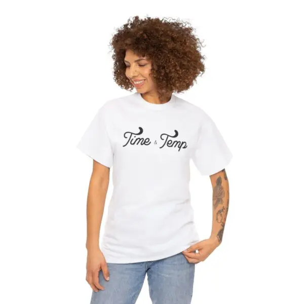 A person with curly hair is wearing a white T-shirt named "Time & Temp Tee," featuring black text that reads "Time & Temp." They are glancing downward with a gentle, effortless smile and have a visible tattoo on their left arm. The background is a plain white.