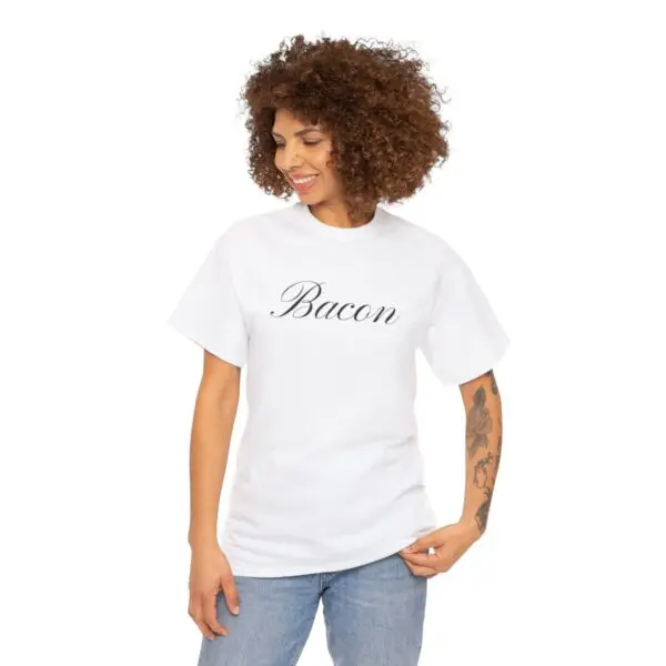 A person with curly hair smiles while wearing the Bacon T-shirt, featuring the word "Bacon" in cursive. They are standing against a plain white background, showcasing their beautiful floral tattoo on their left forearm.