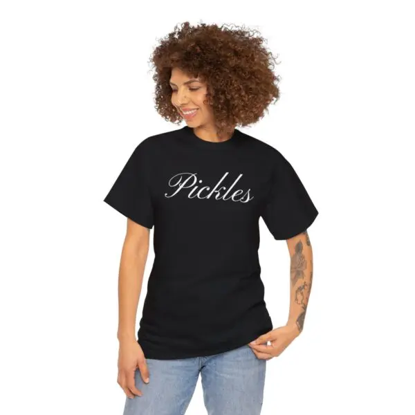 A person with curly hair smiles while wearing the Pickles T-shirt, featuring white script on a black background. They are standing and gazing downward, one hand resting on their hip. Their tattoos lend an effortless charm to their appearance, creating a picture-perfect moment.