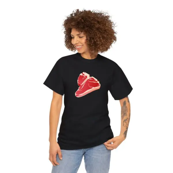 A person with curly hair is wearing a black "Steak T Shirt" that showcases an illustration of a delicious meat cut on the front. They are looking down and smiling contentedly, with one hand gently touching the side of the shirt. Their outfit is completed with blue jeans.