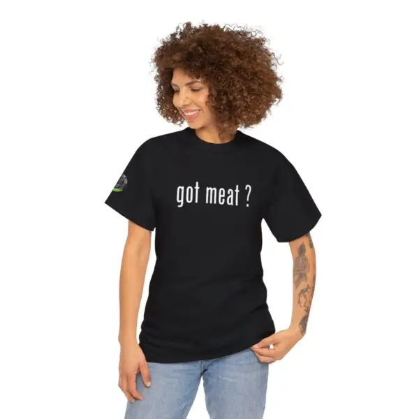 A person with curly hair is wearing a black got meat ? t-shirt, standing casually with one hand pointing to the text. They are also wearing light blue jeans and have eye-catching tattoos on one arm.