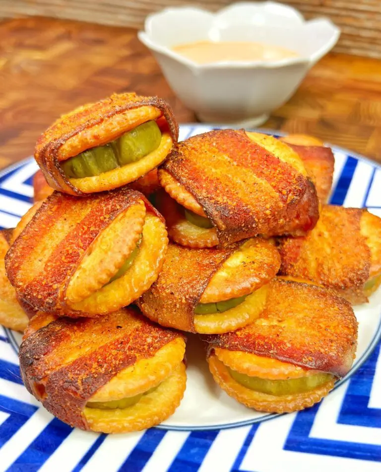 A plate stacked with golden-brown mini sandwiches of pickles wrapped in bacon and seasoned. In the background, a small, white bowl contains a creamy dipping sauce. The plate sits on a blue and white patterned surface.