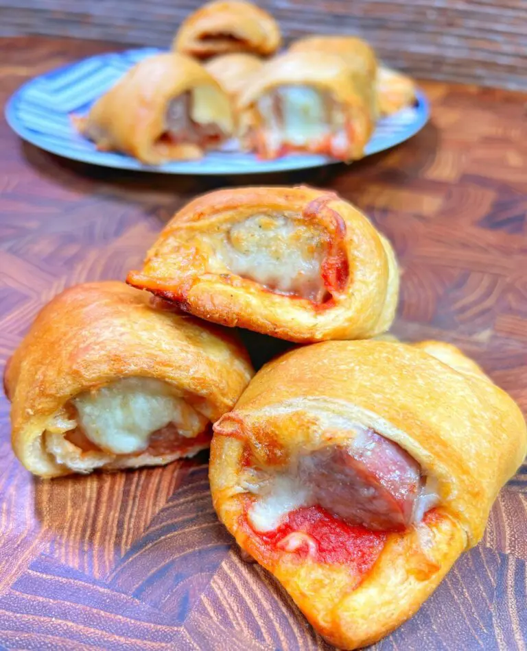 Close-up of golden-brown pizza rolls, reminiscent of Pizza Pigs in a Blanket, filled with melted cheese and pepperoni, resting on a wooden surface. In the background, more rolls are visible on a patterned plate.