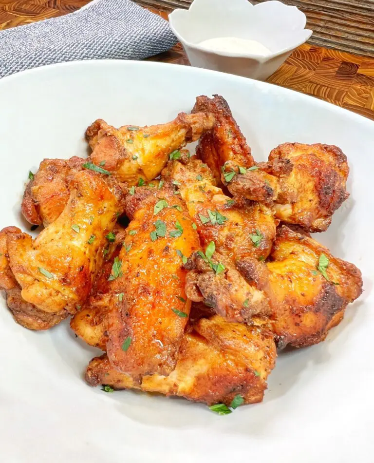 A white bowl filled with crispy, golden-brown chicken wings garnished with chopped herbs—a perfect, tasty delight. In the background, there's an easy creamy dipping sauce in a small white bowl. A textured cloth napkin is partially visible beside it.