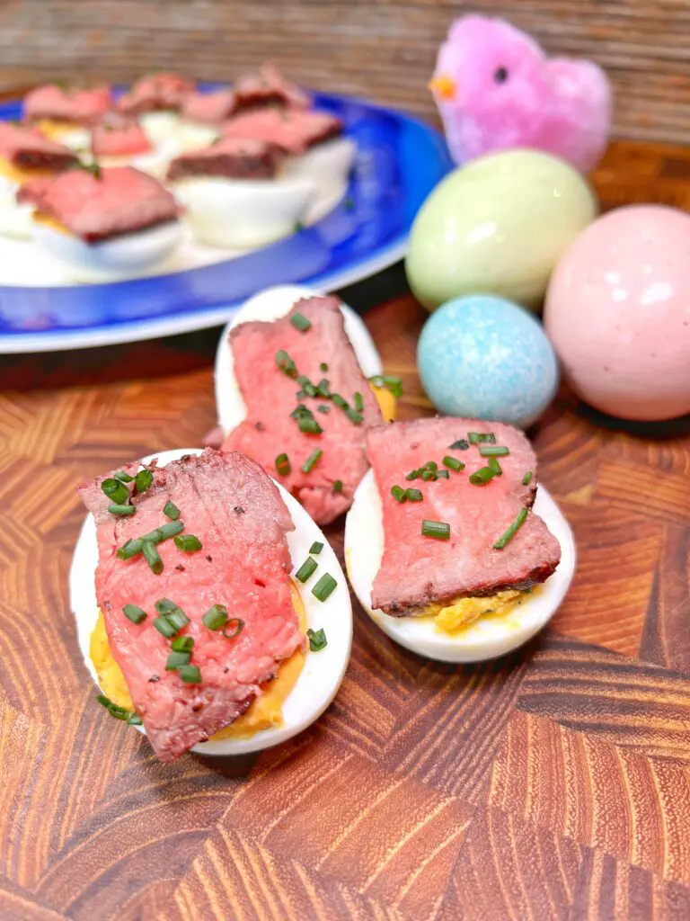 Deviled eggs topped with slices of roast beef and chives sit on a wooden surface. In the background, a blue plate holds more eggs, accompanied by pastel-colored eggs and a small pink chick decoration.