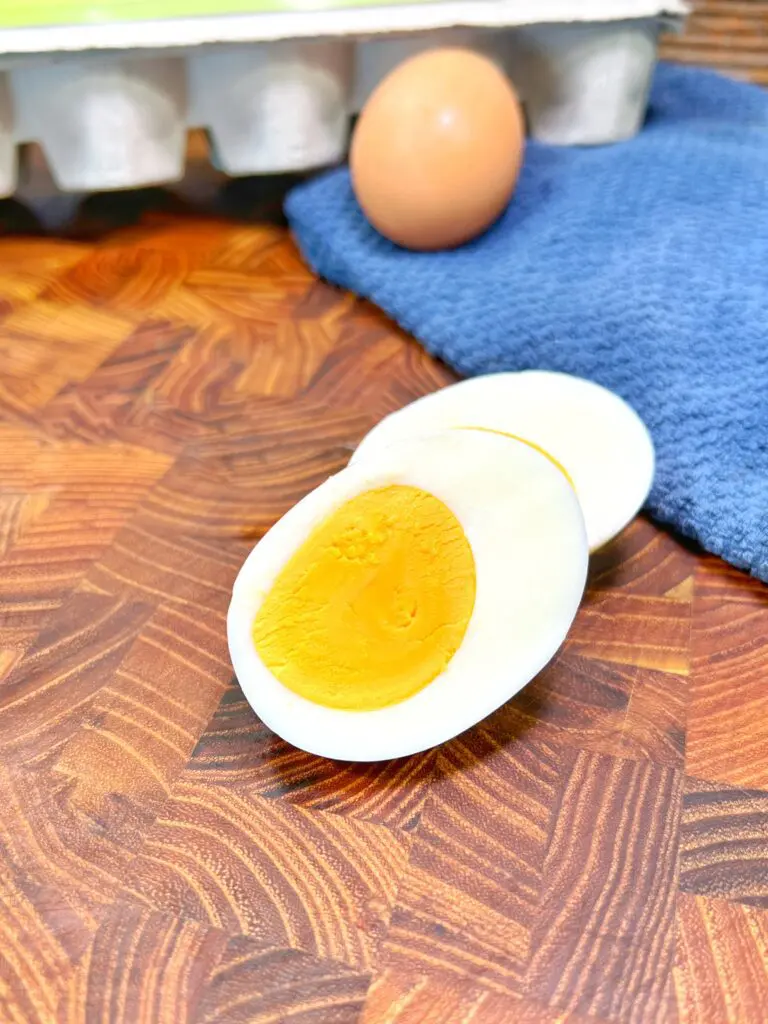 A halved hard-boiled egg with a bright yellow yolk sits perfectly on a wooden surface. In the background, a whole egg rests on a blue cloth, with an egg carton partially visible, hinting at how easy it is to enjoy this delicious treat.