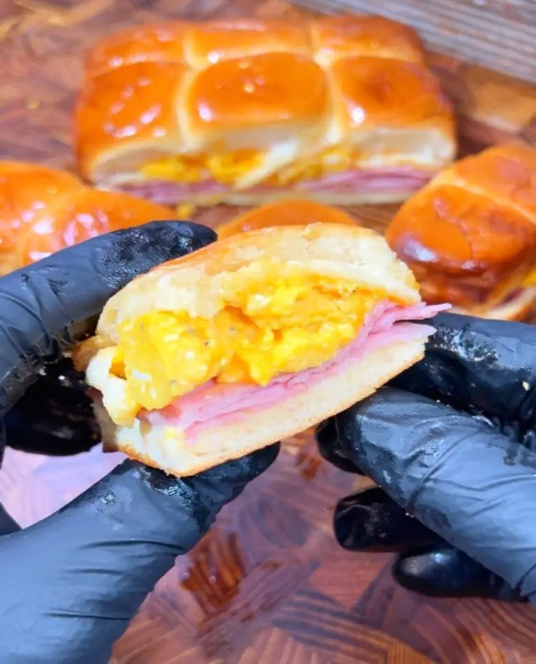 A person wearing black gloves holds a delicious sandwich slider filled with scrambled eggs and ham. More tasty sliders are in the background on a wooden surface. The bread is soft and golden brown.