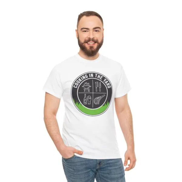 A smiling man with short hair and a beard is wearing the Cooking In The Yard T-Shirt, which showcases illustrations of a grill, spatula, matchstick, and leaf. He stands against a plain white background, embodying the spirit of delicious outdoor cooking.