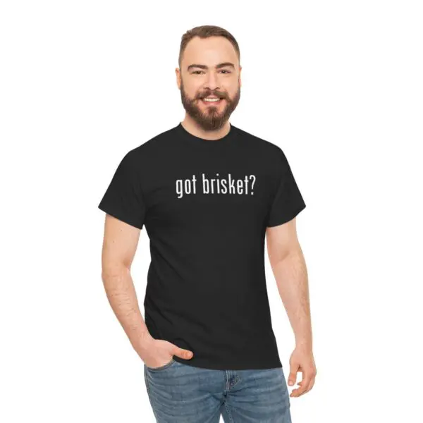 A bearded, smiling man wearing a black T-shirt featuring the "got brisket ?" slogan stands against a white background. With one hand in the pocket of his blue jeans, he perfectly captures an easygoing style that complements the deliciously quirky message on his shirt.