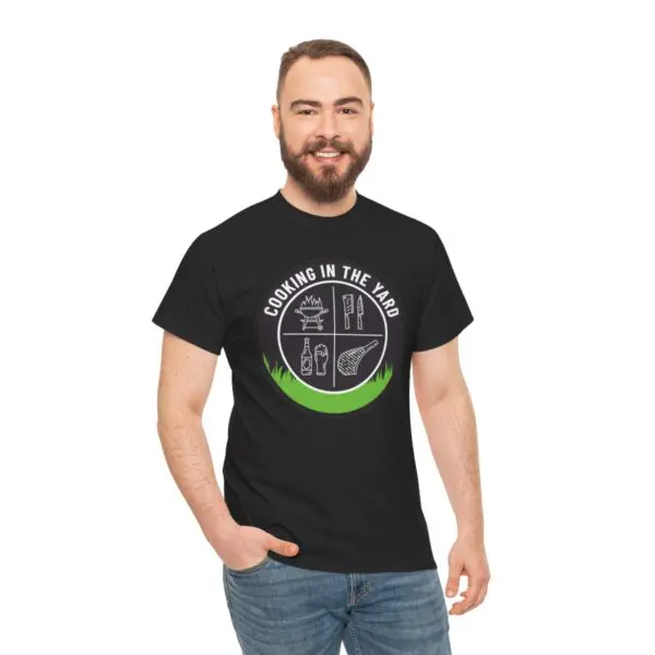 A cheerful man with a beard dons the Cooking In The Yard T-Shirt, featuring illustrations of outdoor cooking utensils in black. He stands against a simple white backdrop, exuding effortless charm with one hand tucked into his jeans pocket, poised to create something delectable.