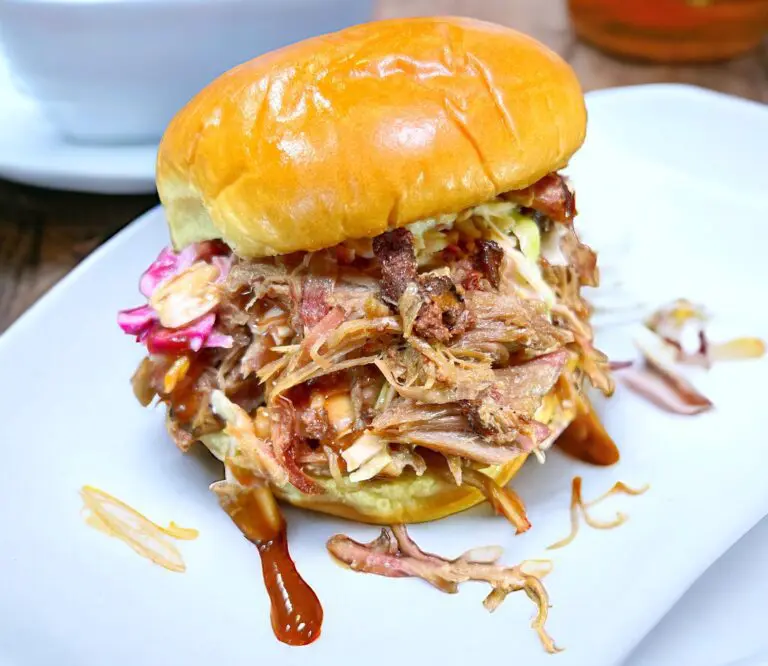 A delicious pulled pork sandwich sits on a white plate, topped with coleslaw and a glossy brioche bun. Tasty barbecue sauce drips from the sides, with a splash artfully decorating the plate.