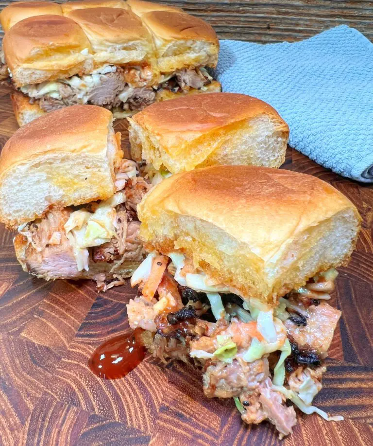 Pulled pork sliders with coleslaw and BBQ sauce on wooden board, next to a blue cloth napkin.