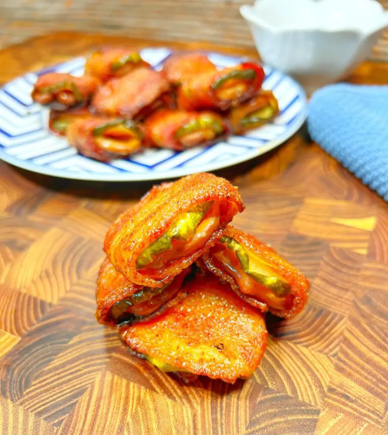 Close-up of crispy bacon-wrapped jalapeño poppers stacked on a wooden surface. A plate with more poppers is blurred in the background, alongside a folded blue napkin and a small white sauce dish.