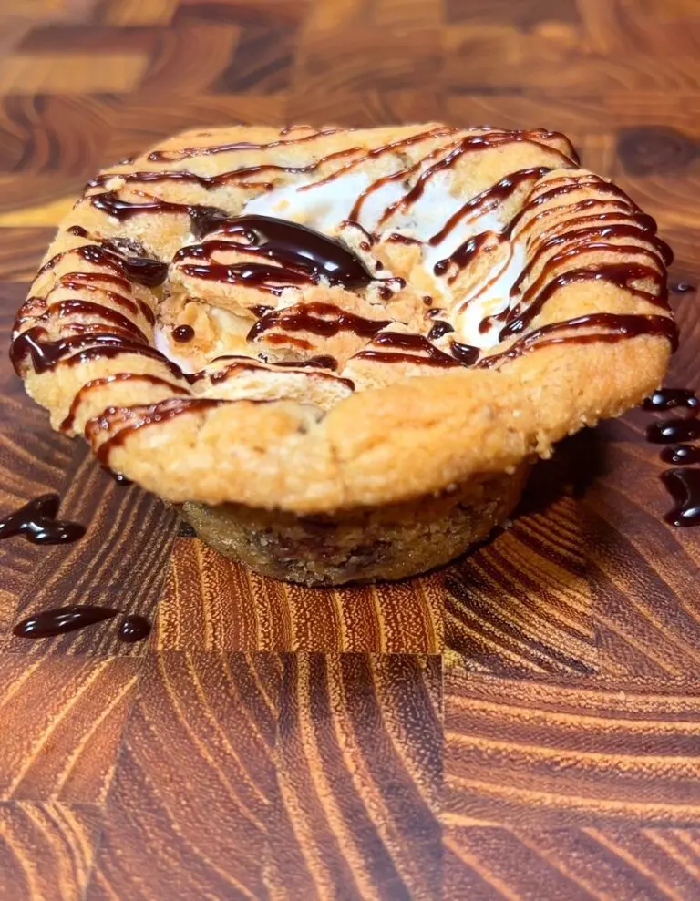 A close-up of a cookie cup filled with gooey marshmallow and drizzled with chocolate sauce, placed on a wooden surface with rich grain patterns.