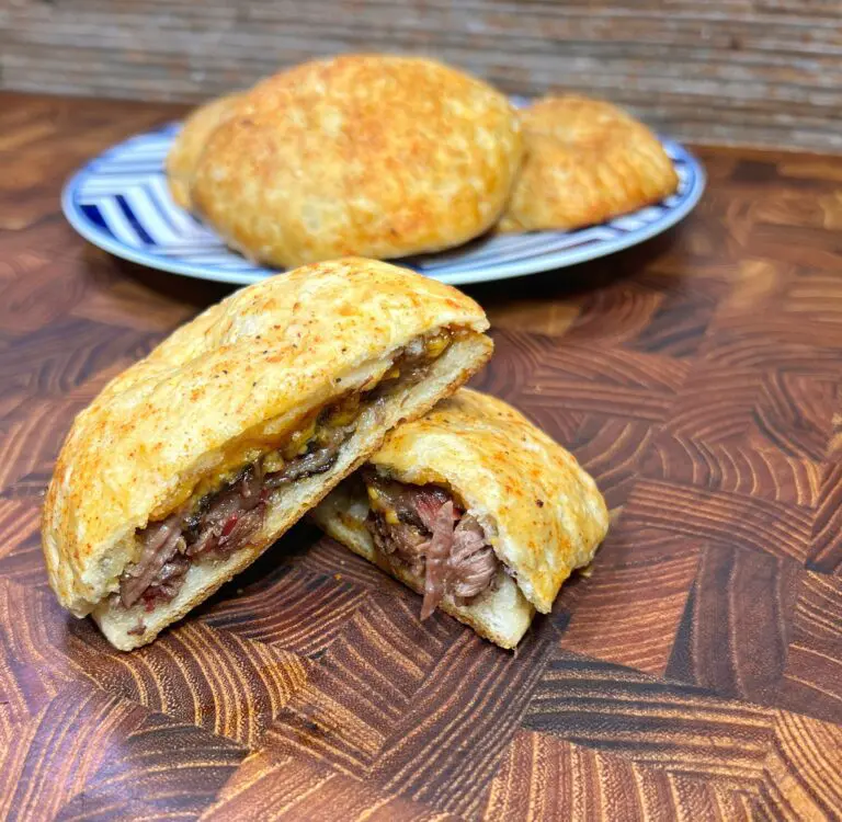 A close-up of a sliced baked pastry filled with shredded meat on a wooden surface. In the background, a plate holds whole pastries. The scene highlights the texture of the flaky crust and savory filling.