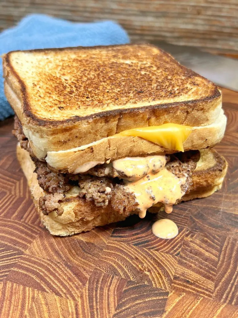 A grilled cheese sandwich with a beef patty and melted cheese sauce, sandwiched between two slices of toasted bread. It is placed on a wooden surface, with a blue cloth in the background.