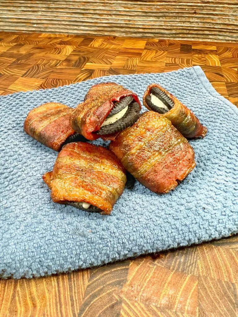 Delicious bacon-wrapped cookies rest on a blue towel atop a wooden surface. The partially cut cookies reveal a creamy filling inside, making them irresistibly tasty.