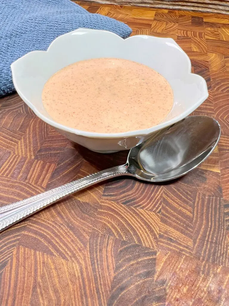 A white scalloped bowl filled with creamy tomato soup rests on a wooden table beside a large silver spoon. A blue cloth is partially visible in the background.