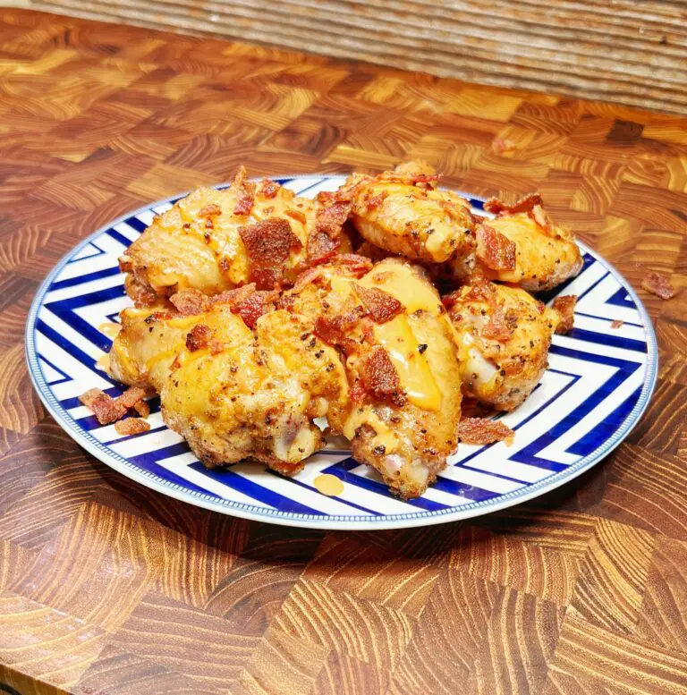 A plate of crispy baked chicken wings, topped with bacon bits and drizzled with honey, offers a perfectly tasty treat. Served on a blue and white patterned plate, the dish rests invitingly on a wooden table against a textured background.