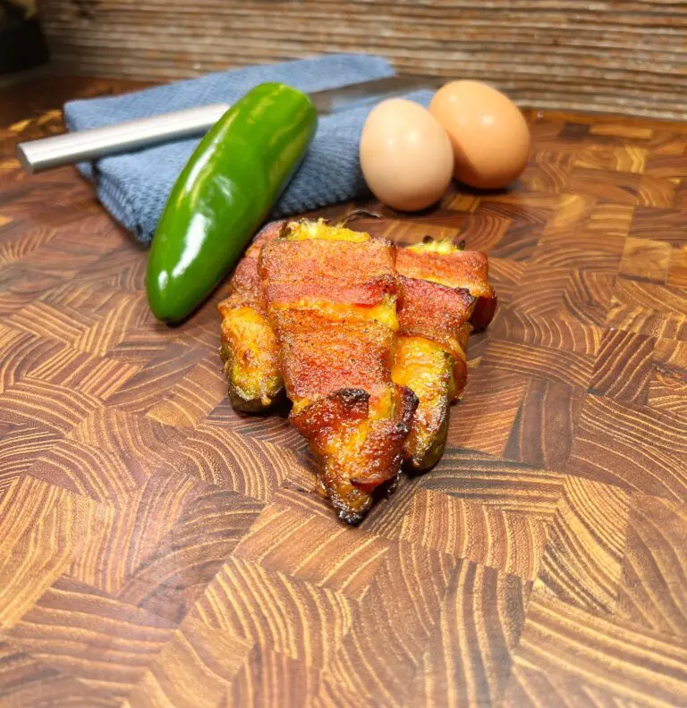 A stack of bacon-wrapped jalapeños on a wooden countertop. In the background, there is a whole jalapeño, two eggs, and a blue cloth next to a silver knife. The surface has a rich wood grain pattern.