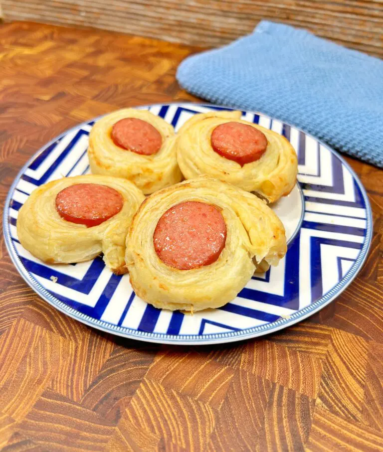 A plate of spiral puff pastries with slices of sausage on top, placed on a blue and white zigzag-patterned plate. The background is a wooden surface with a folded blue cloth on the side.