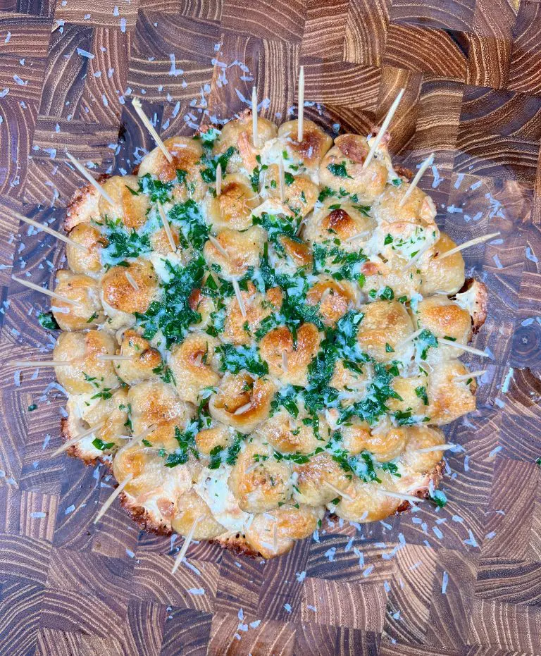 A pull-apart garlic bread flower topped with melted cheese and chopped parsley, served on a wooden board. Small skewers are inserted for easy serving.