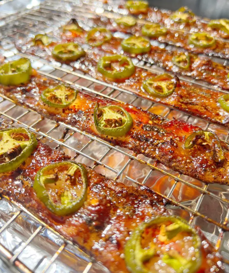 Slices of glazed bacon topped with jalapeño slices rest on a wire rack, glistening in the sunlight.