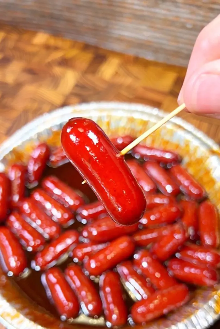 A hand holds a mini sausage on a toothpick above a tray filled with more sausages in a glossy sauce. The sausages are shiny and dark red, suggesting they are cooked in a rich glaze.