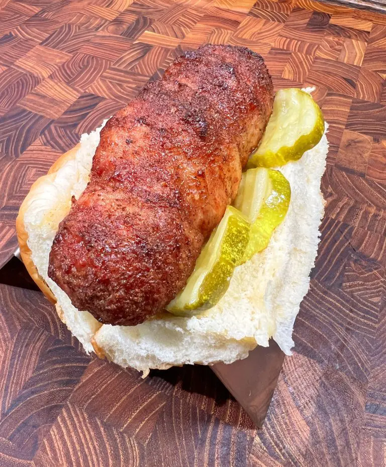 Grilled meatloaf topped with pickles, served in a hot dog bun on a wooden cutting board.