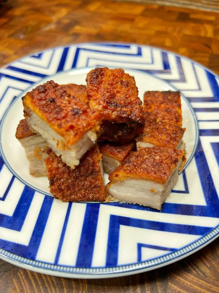 A plate with several pieces of crispy, golden-brown pork belly, stacked and arranged neatly. The plate has a blue and white geometric pattern, placed on a wooden table.