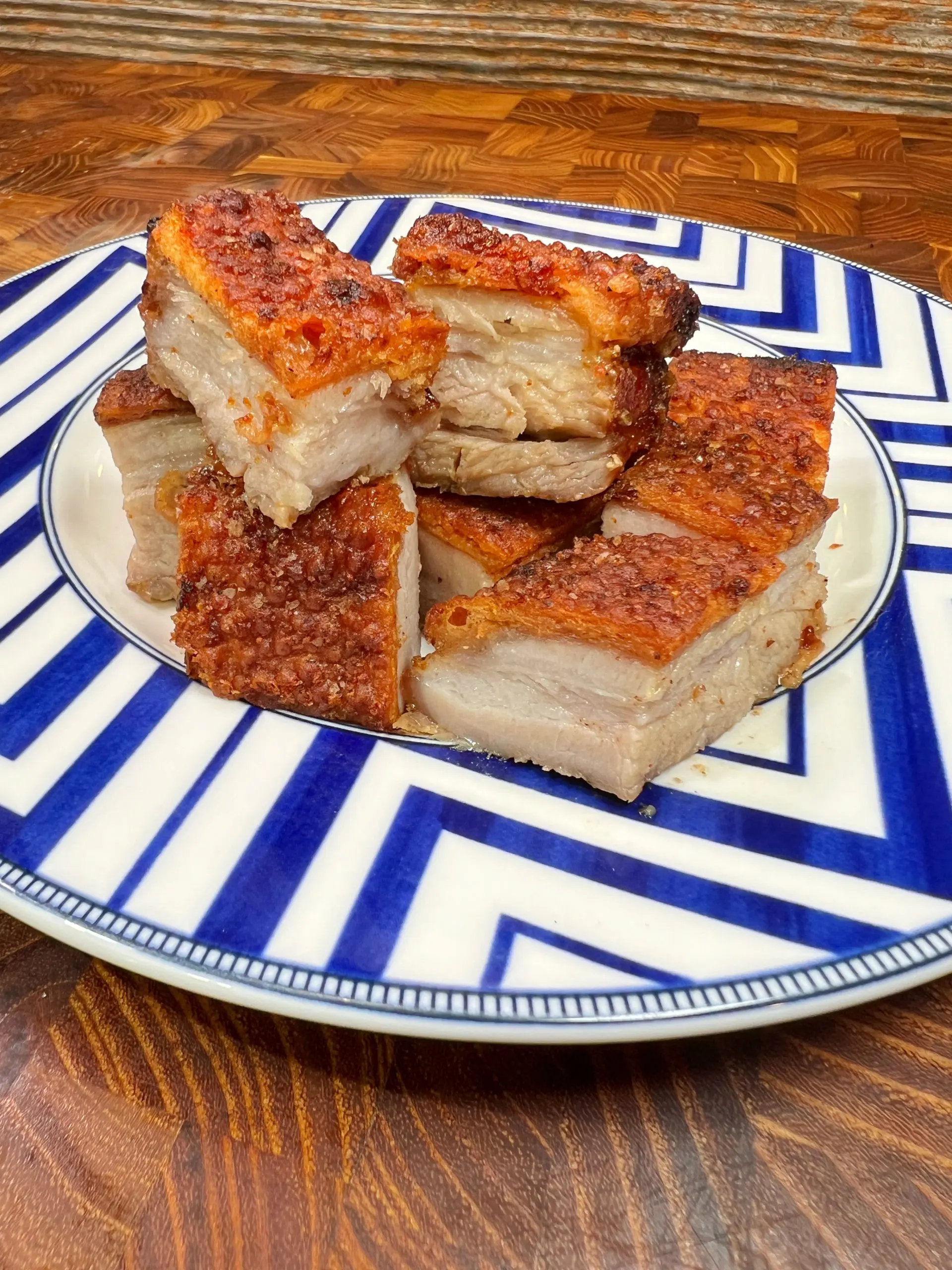 A plate of crispy roast pork with golden-brown skin, cut into rectangular pieces, is arranged on a white plate with blue geometric patterns. The plate is set on a wooden table.