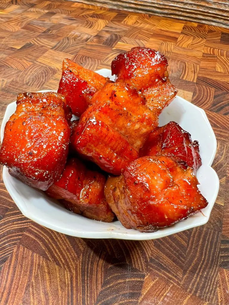 A white bowl filled with glossy, caramelized cubes of roasted pork, resting on a wooden textured surface. The pork pieces are glazed, giving them a rich, appetizing appearance.