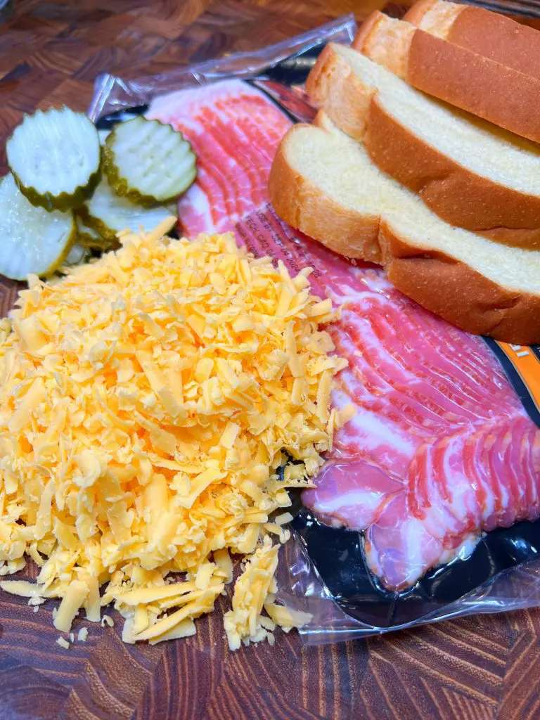 Ingredients for grilled cheese with pickles sandwich are on a wooden table: sliced bread, thin bacon strips, shredded cheddar cheese, and pickle slices.