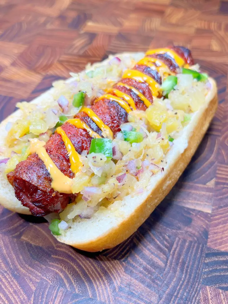 A grilled sausage in a bun topped with diced onions, green peppers, pineapple relish, and drizzled mustard. The hot dog is served on a wooden surface.