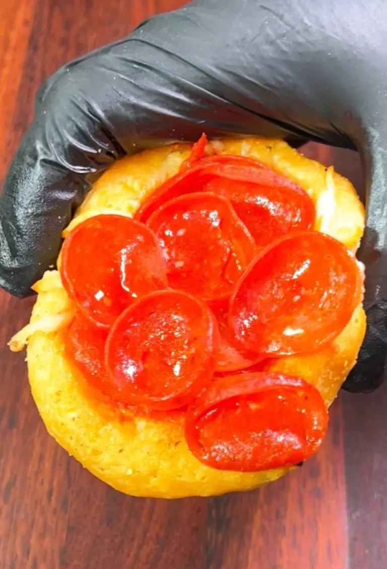 A hand in a black glove holding a donut topped with several slices of pepperoni on a wooden surface.