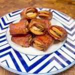 A plate of pecan-stuffed dates wrapped in crispy bacon, served on a blue and white zigzag-patterned dish. The wooden background adds warmth to the presentation.