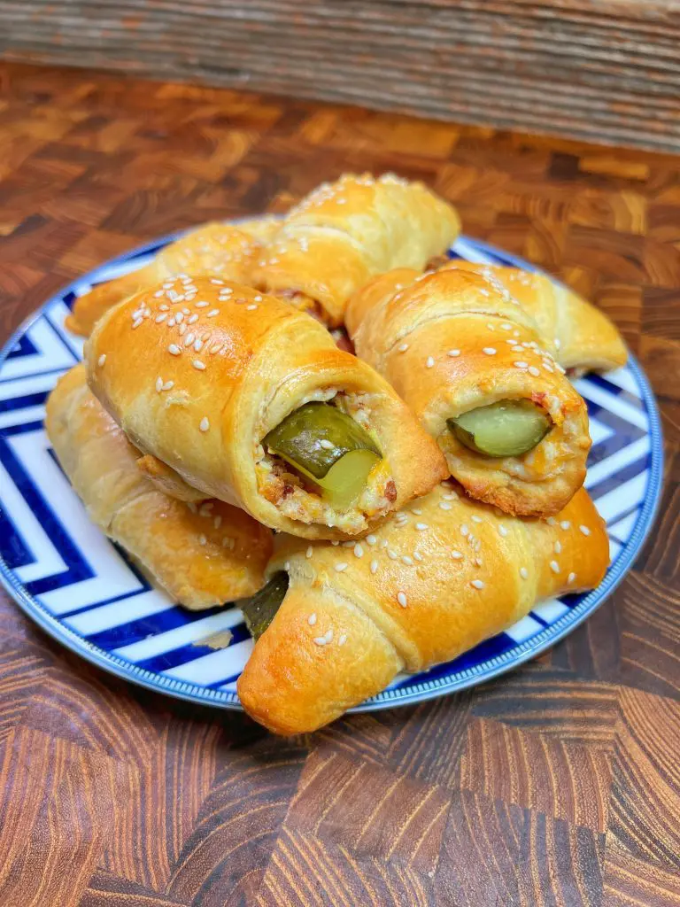 A plate of freshly baked, golden-brown crescent rolls filled with pickles and sprinkled with sesame seeds. They rest on a blue and white patterned plate, placed on a wooden surface.