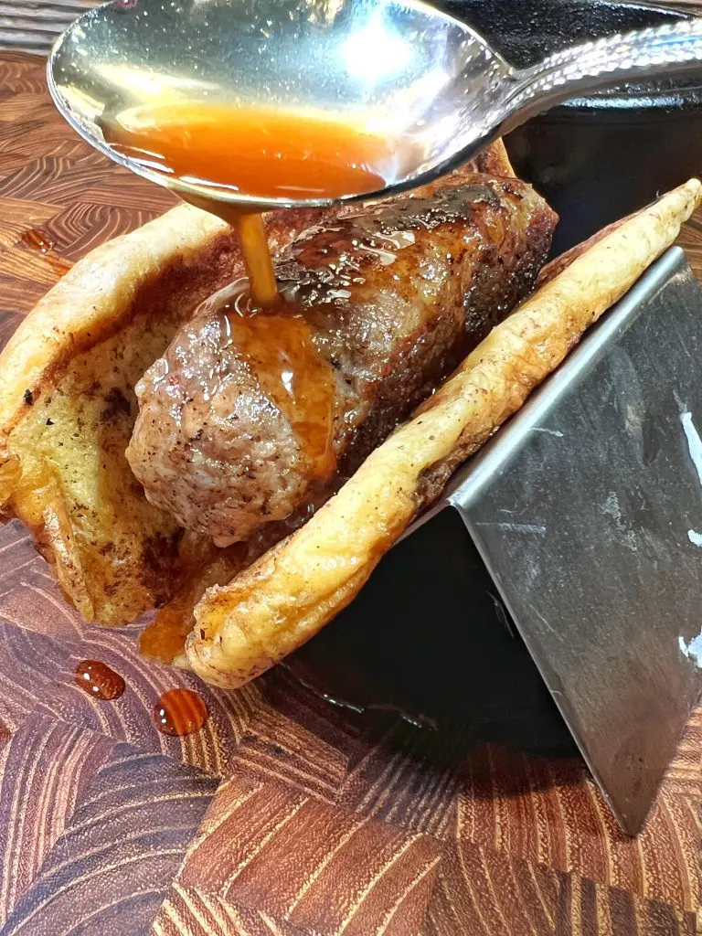 A grilled sausage resting inside flaky pastry is being drizzled with sauce from a spoon. The ensemble is supported on a black stand, set against a textured wooden surface.