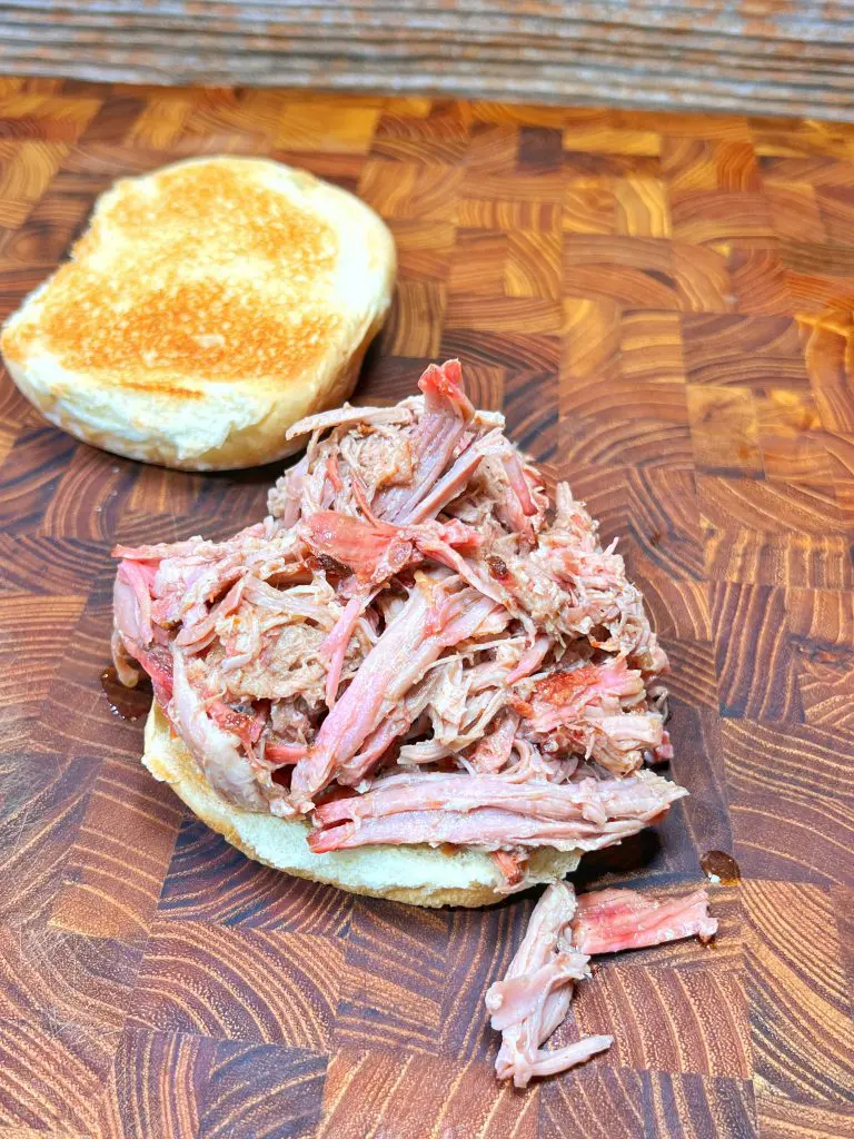 A sandwich bun with a generous serving of delicious shredded pulled pork rests on a wooden surface. The toasted top half of the bun is positioned at the back, making this tasty treat easy to enjoy.