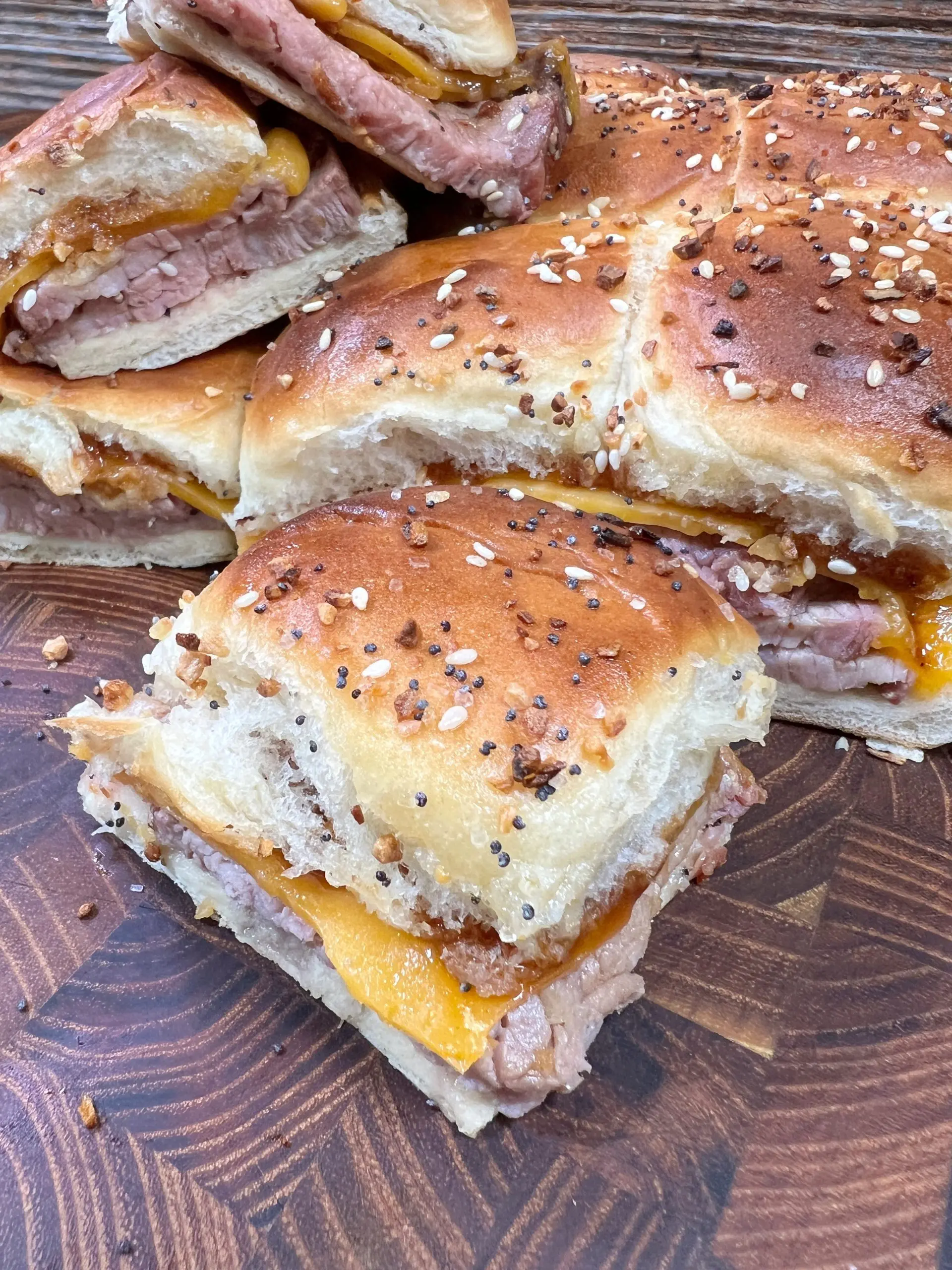 A close-up of sliced sandwiches on a wooden board. The sandwiches contain layers of beef and melted cheese, topped with a sprinkled seed seasoning on golden-brown buns.